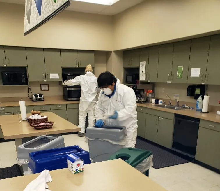 Certified decontamination specialists wearing full protective gear performing sanitization in a breakroom.